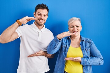 Sticker - Young brazilian mother and son standing over blue background gesturing with hands showing big and large size sign, measure symbol. smiling looking at the camera. measuring concept.