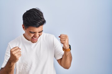 Poster - Hispanic man standing over blue background very happy and excited doing winner gesture with arms raised, smiling and screaming for success. celebration concept.