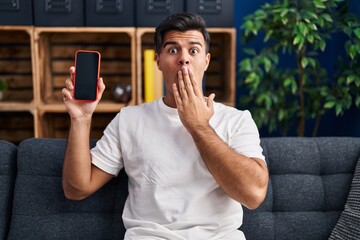 Sticker - Hispanic man holding smartphone showing blank screen covering mouth with hand, shocked and afraid for mistake. surprised expression