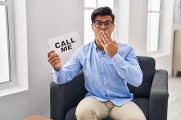 Canvas Print - Hispanic man working at therapy office holding call me banner covering mouth with hand, shocked and afraid for mistake. surprised expression