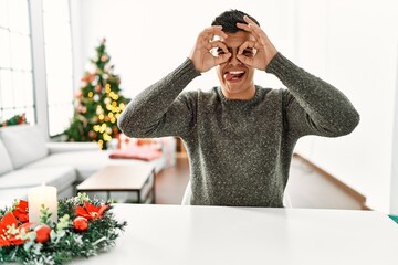 Canvas Print - Young hispanic man sitting on the table by christmas tree doing ok gesture like binoculars sticking tongue out, eyes looking through fingers. crazy expression.