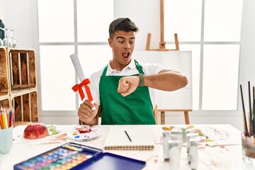 Poster - Young hispanic man at art studio holding degree looking at the watch time worried, afraid of getting late