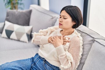 Sticker - Young chinese woman sitting on sofa with hands on heart at home