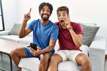 Poster - Young hispanic men using smartphone sitting on the sofa at home pointing finger up with successful idea. exited and happy. number one.