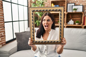 Sticker - Young hispanic woman holding empty frame angry and mad screaming frustrated and furious, shouting with anger. rage and aggressive concept.