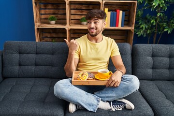 Canvas Print - Arab man with beard sitting on the sofa at home eating breakfast pointing thumb up to the side smiling happy with open mouth