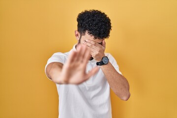 Sticker - Arab man standing over yellow background covering eyes with hands and doing stop gesture with sad and fear expression. embarrassed and negative concept.