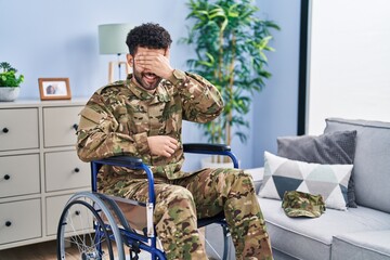 Poster - Arab man wearing camouflage army uniform sitting on wheelchair smiling and laughing with hand on face covering eyes for surprise. blind concept.