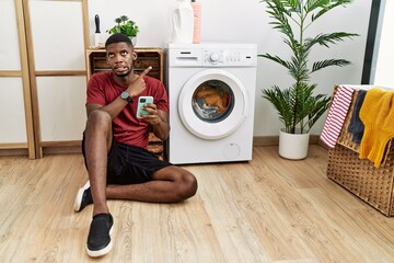 Canvas Print - Young african american man using smartphone waiting for washing machine pointing aside worried and nervous with forefinger, concerned and surprised expression