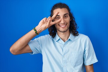 Poster - Young hispanic man standing over blue background doing peace symbol with fingers over face, smiling cheerful showing victory