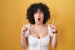 © Krakenimages.com - Young brunette woman with curly hair standing over yellow background amazed and surprised looking up and pointing with fingers and raised arms.