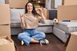 © Krakenimages.com - Young brunette woman sitting on the floor at new home holding keys smiling happy pointing with hand and finger to the side