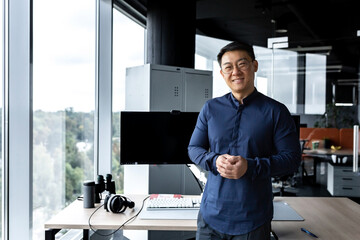 Programmer in glasses and shirt smiling and looking at camera, Asian man working inside modern computer company, working in office with several computers and monitors.