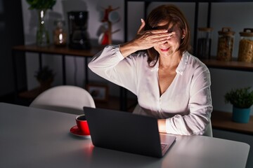 Poster - Middle age hispanic woman using laptop at home at night smiling and laughing with hand on face covering eyes for surprise. blind concept.