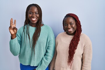 Sticker - Two african woman standing over blue background showing and pointing up with fingers number three while smiling confident and happy.