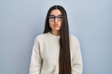 Poster - Young hispanic woman wearing casual sweater over blue background puffing cheeks with funny face. mouth inflated with air, crazy expression.