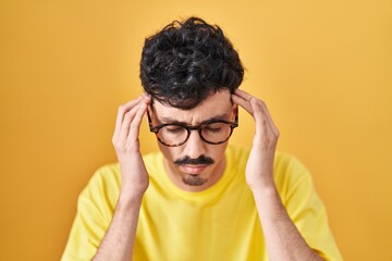 Hispanic man wearing glasses standing over yellow background with hand on head for pain in head because stress. suffering migraine.