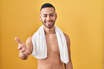 Wall Mural - Young hispanic man standing shirtless with towel smiling cheerful offering palm hand giving assistance and acceptance.