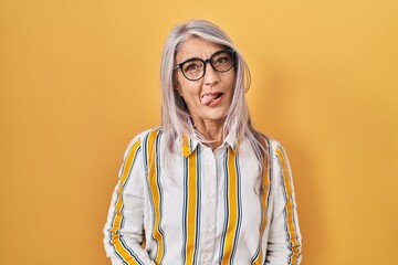 Wall Mural - Middle age woman with grey hair standing over yellow background wearing glasses sticking tongue out happy with funny expression. emotion concept.