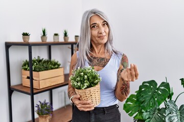 Sticker - Middle age grey-haired woman holding green plant pot at home doing money gesture with hands, asking for salary payment, millionaire business