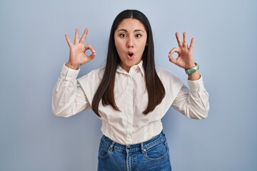 Wall Mural - Young latin woman standing over blue background looking surprised and shocked doing ok approval symbol with fingers. crazy expression