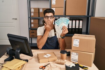 Canvas Print - Young hispanic man working at small business ecommerce holding brazilian reals covering mouth with hand, shocked and afraid for mistake. surprised expression