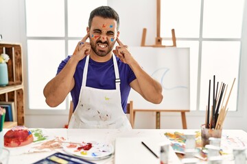 Poster - Young hispanic man with beard at art studio with painted face covering ears with fingers with annoyed expression for the noise of loud music. deaf concept.