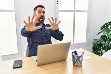 Poster - Young hispanic man with beard working at the office with laptop afraid and terrified with fear expression stop gesture with hands, shouting in shock. panic concept.