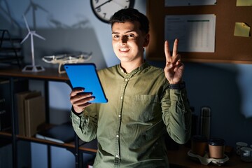 Canvas Print - Non binary person using touchpad device at night smiling with happy face winking at the camera doing victory sign with fingers. number two.