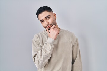 Canvas Print - Young handsome man standing over isolated background looking confident at the camera smiling with crossed arms and hand raised on chin. thinking positive.