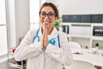 Canvas Print - Young brunette doctor woman wearing stethoscope at the clinic praying with hands together asking for forgiveness smiling confident.