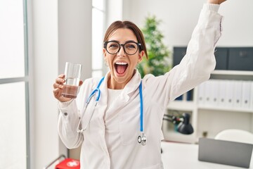 Wall Mural - Young brunette doctor woman holding glass of water celebrating victory with happy smile and winner expression with raised hands