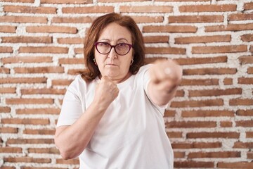 Poster - Senior woman with glasses standing over bricks wall punching fist to fight, aggressive and angry attack, threat and violence