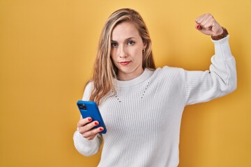 Wall Mural - Young blonde woman using smartphone typing message strong person showing arm muscle, confident and proud of power