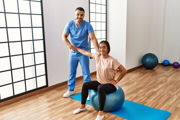 Wall Mural - Latin man and woman wearing physiotherapist uniform having rehab session using fit ball at rehab center