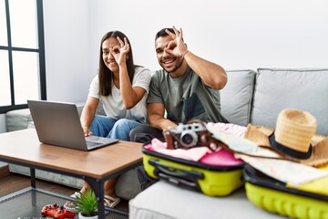 Canvas Print - Young interracial couple packing summer clothes in suitcase looking at laptop smiling happy doing ok sign with hand on eye looking through fingers