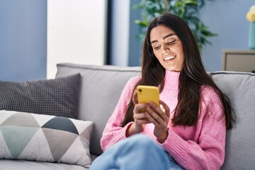 Wall Mural - Young hispanic woman using smartphone sitting on sofa at home