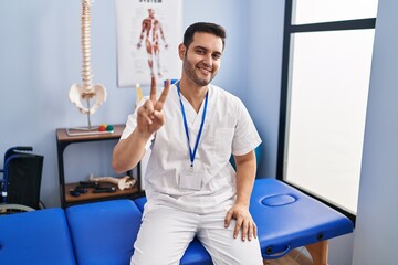 Sticker - Young hispanic man with beard working at pain recovery clinic smiling looking to the camera showing fingers doing victory sign. number two.