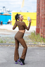 Pretty Asian Woman Wearing Tight Fitting Two Piece Checkered Bodysuit And Rollerskates. She Is Looking Back Over Her Shoulder And Giving A Peace Sign Gesture With Her Fingers. 