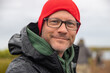 © Mat Hayward - Close up face of handsome man wearing red beanie hat and glasses. He has beard stubble and a friendly smile.