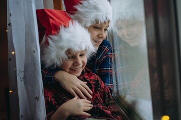  cute caucasian children wearing snta hats and plaid shirts sitting hugging by the window waitinf for christmas