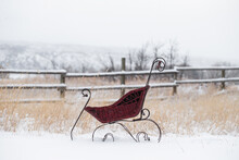 Antique Baby Sled Pram In Snowy Winter Field As It Is Snowing With An Old Primitive Wooden Fence Sparse Tall Grasses And Bare Trees In The Background For Photography Or Digital Artist Composite Layer