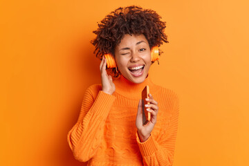 Wall Mural - Happy curly haired woman listens audio track via headphones holds smartphone as if microphone near mouth winks eyes has glad expression foolishes around wears casual jumper isolated over orange wall