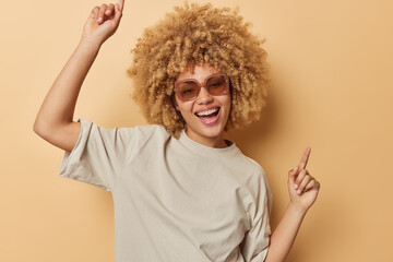 Wall Mural - Carefree optimistic woman with curly hair shakes arms dances has fun smiles broadly wears sunglasses and casual t shirt isolated on beige studio background. Cheerful female model being full of energy