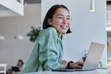 Fototapeta  - Pretty cheerful Asian woman in eyeglasses and casual clothes browses laptop computer connected to 4g internet updates software uses modern technologies poses in cafeteria looks gladfully away