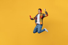 Full Body Young Fun Happy Cool Middle Eastern Man Wears Casual Shirt White T-shirt Headphones Listen Music Jump High Do Winner Gesture Hold Use Mobile Cell Phone Isolated On Plain Yellow Background.