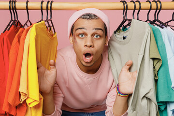 Wall Mural - Young fun amazed gay man wear sweatshirt hat choose basic colorful collection of t-shirts on hangers in shop isolated on plain pastel light pink color background studio. Lifestyle lgbtq pride concept.