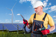 Construction of solar power plants. Man holds tablet and screwdriver. Guy is building power plants. Solar panels and wind turbines under blue sky. Male engineer on field with ECO power plant.