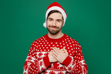 Merry thankful young man 20s wear red warm knitted sweater Santa hat posing put folded hands on heart isolated on plain dark green background studio portrait New Year 2023 holiday celebration concept