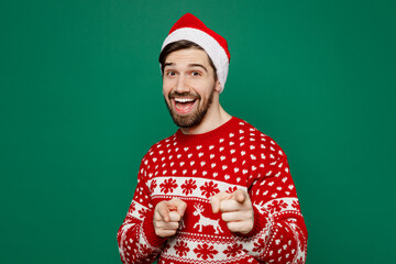 Wall Mural - Merry young man wear red warm knitted sweater Santa hat posing point index finger camera on you motivating encourage isolated on plain dark green background. New Year 2023 holiday celebration concept.